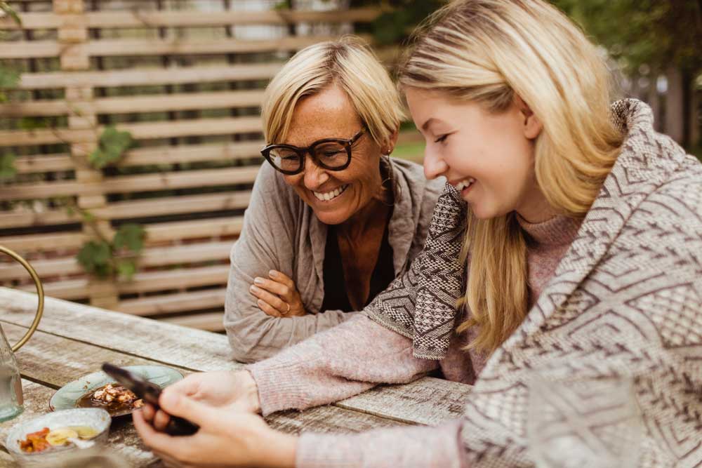 Äldre mamma sitter och tittar på något i mobilen tillsammans med sin dotter.